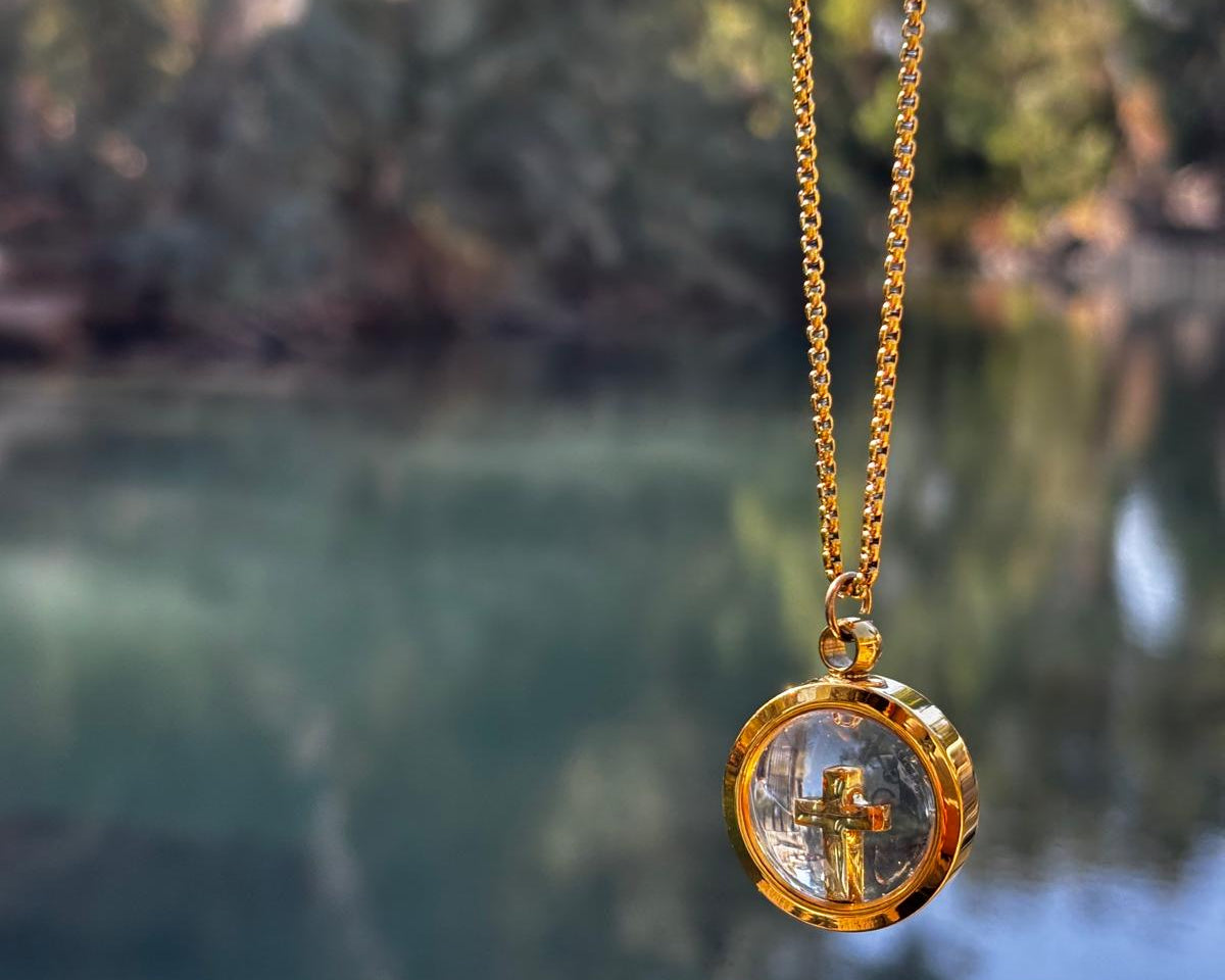 Gold necklace with a clear pendant featuring a cross, against a blurred natural background.